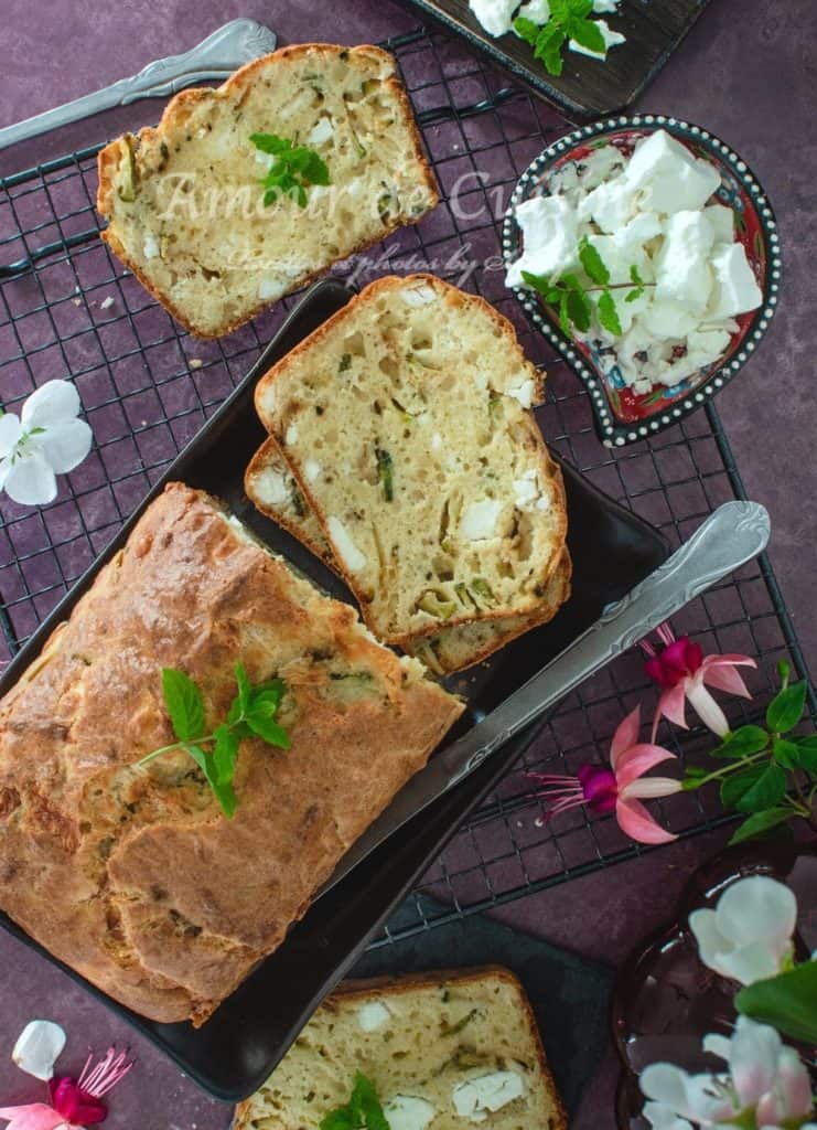 Top view of sliced zucchini and feta savoury cake on a cooling rack, with a block of feta cheese on a decorative plate, garnished with mint and surrounded by fresh flowers