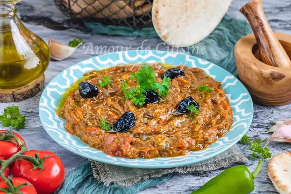 Traditional Moroccan zaalouk served in a turquoise patterned plate, garnished with black olives and parsley, with olive oil, tomatoes, green pepper, garlic, and flatbread in the background