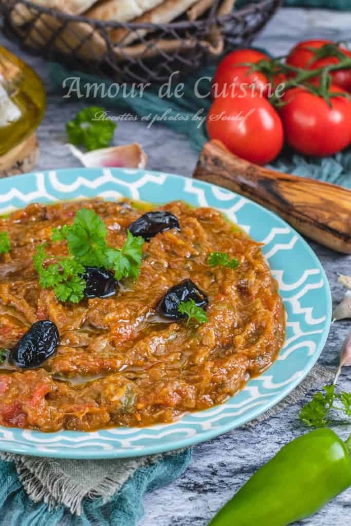 Close-up of Moroccan zaalouk served in a turquoise patterned dish, garnished with black olives and fresh parsley, with tomatoes, garlic, green pepper, and Moroccan flatbread in the background