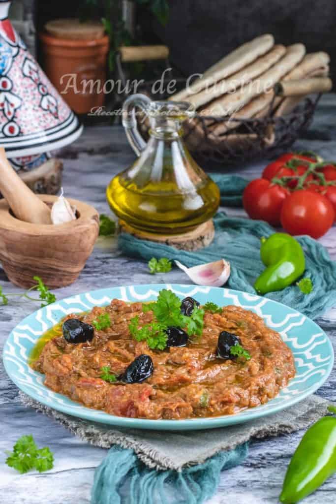 Traditional Moroccan zaalouk served in a patterned turquoise plate, garnished with black olives and fresh parsley, surrounded by tomatoes, green pepper, olive oil, garlic, and Moroccan bread in the background