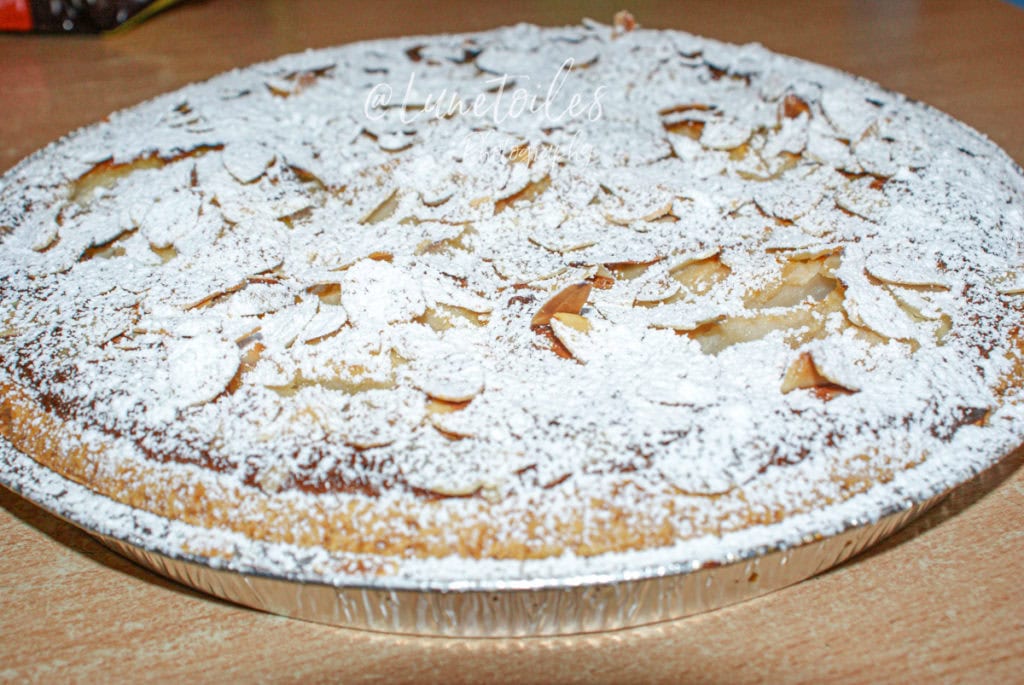 Whole pear amandine tart baked in a round aluminum pan, topped with flaked almonds and generously dusted with powdered sugar, displayed on a wooden surface.