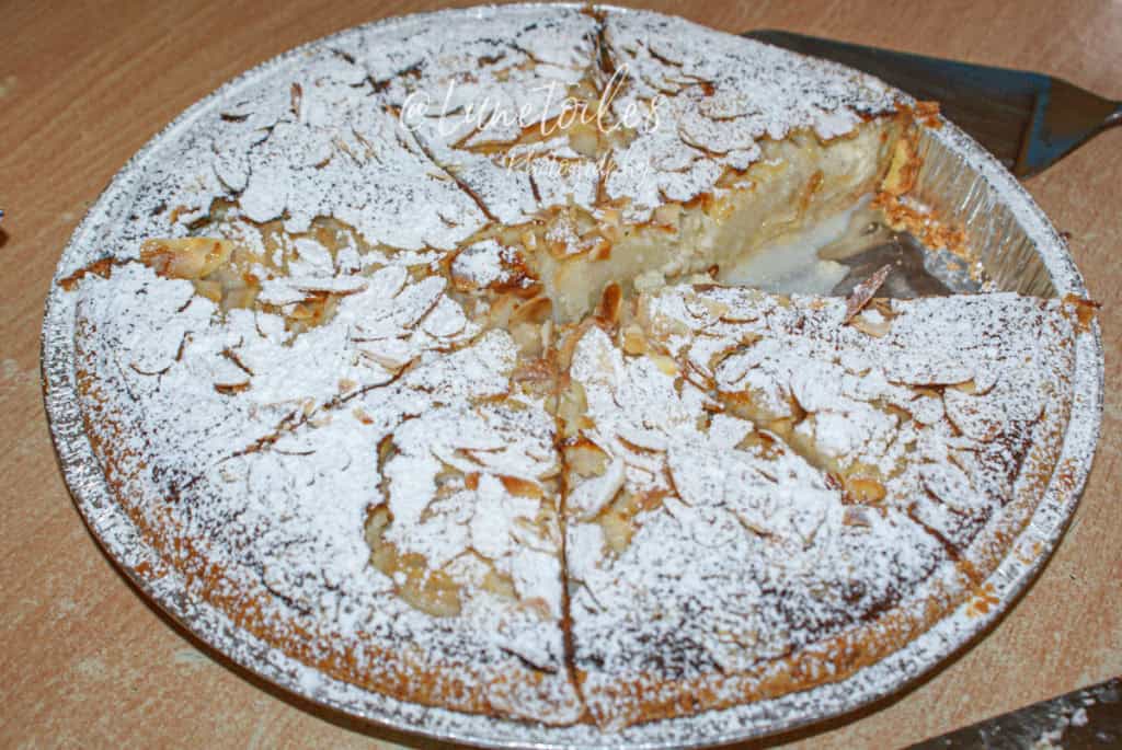 Partially sliced pear amandine tart in a round aluminum pan, topped with flaked almonds and powdered sugar, revealing a creamy almond filling inside