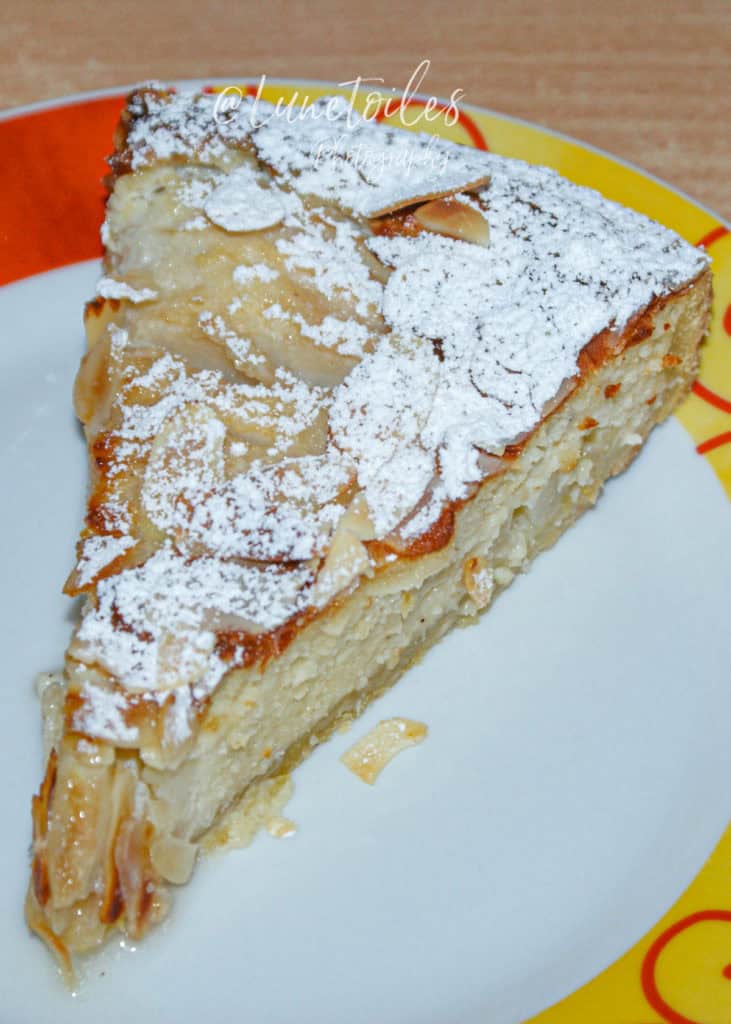 Slice of pear amandine tart topped with flaked almonds and powdered sugar, served on a colorful plate with a golden, moist almond filling visible