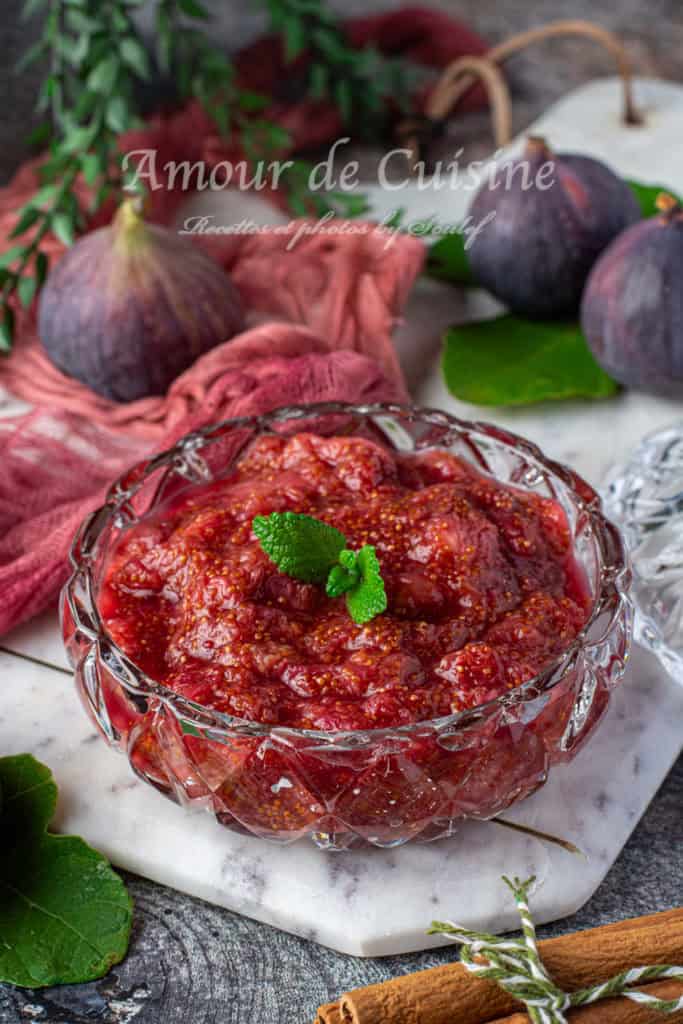 A close-up of a glass bowl filled with sugar-free fig compote, garnished with a fresh mint leaf on top. The compote has a rich, deep red color and a thick texture with visible fig seeds. It is placed on a white marble serving board surrounded by fresh figs, green leaves, a cinnamon stick, and a red cloth, creating a cozy and inviting rustic setting.
