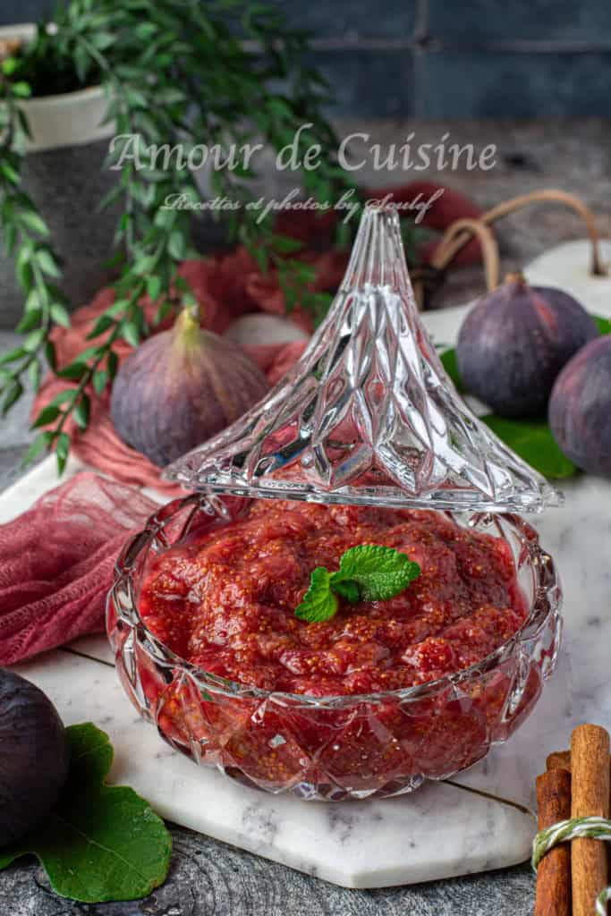 A glass dish with a decorative lid filled with thick, sugar-free fig compote, topped with a fresh mint leaf. The compote has a rich, reddish-purple color with visible fig seeds. It is placed on a white marble slab surrounded by fresh figs, green fig leaves, cinnamon sticks, and a red cloth, creating a warm and rustic presentation. The background includes greenery and a dark stone wall
