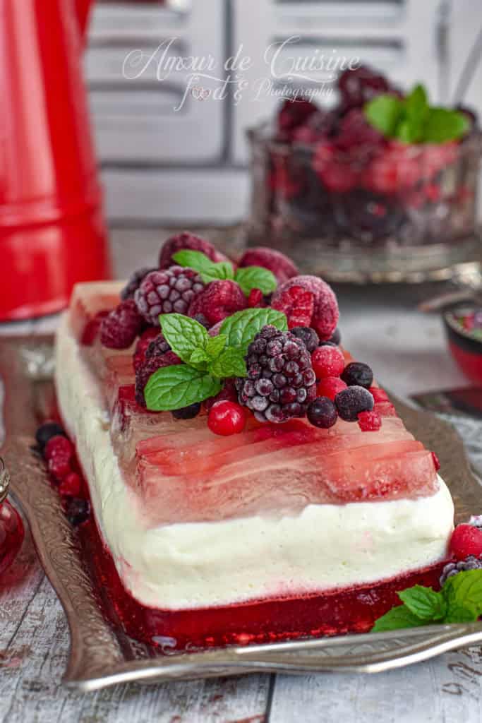 Layered summer berry panna cotta terrine with vanilla cream and red fruit jelly, topped with frozen mixed berries and mint leaves, served on a vintage silver tray