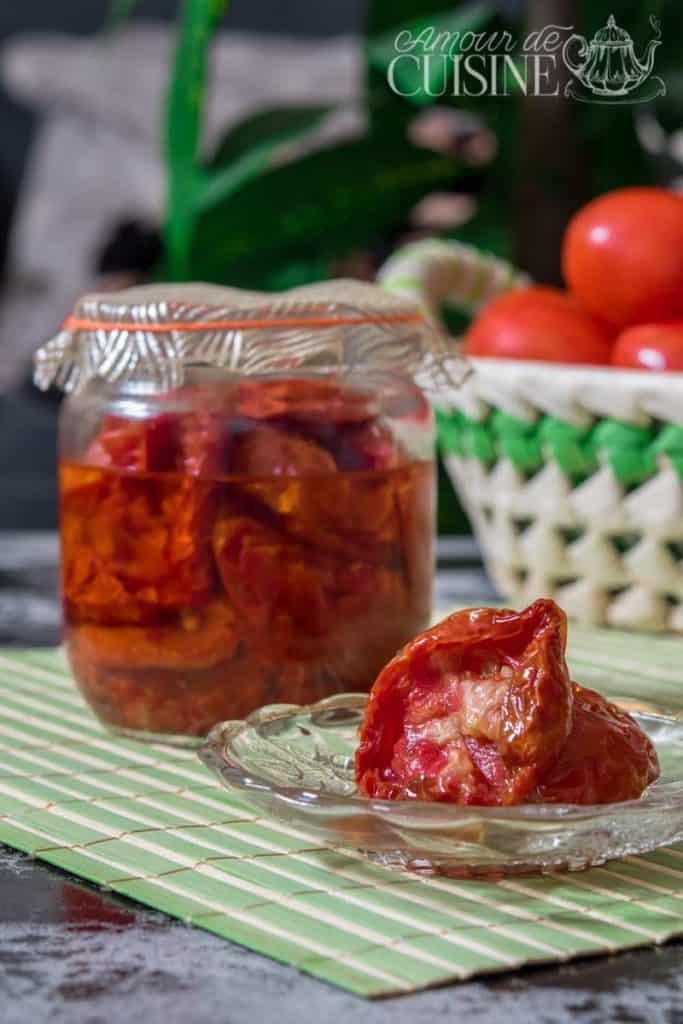 Jar of homemade sun-dried tomatoes in oil with a small glass plate holding a few tomatoes in front, set on a green-striped mat with fresh tomatoes and a woven basket in the background