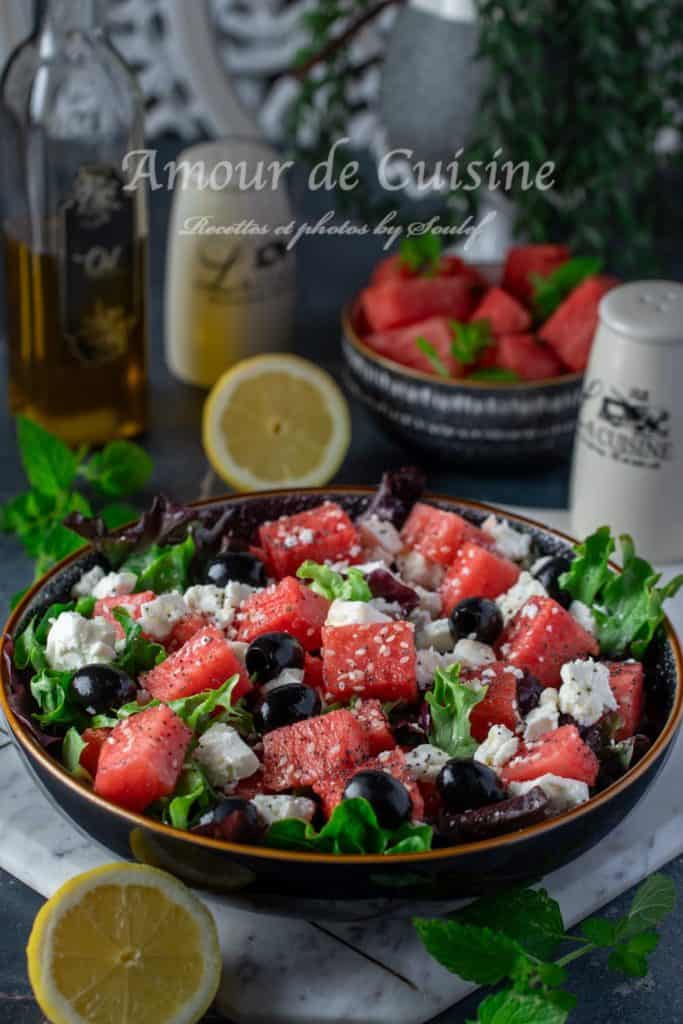 Watermelon feta salad served in a black bowl with black olives, fresh greens, sesame and poppy seeds, surrounded by lemon halves, mint leaves, olive oil, and extra watermelon cubes in the background