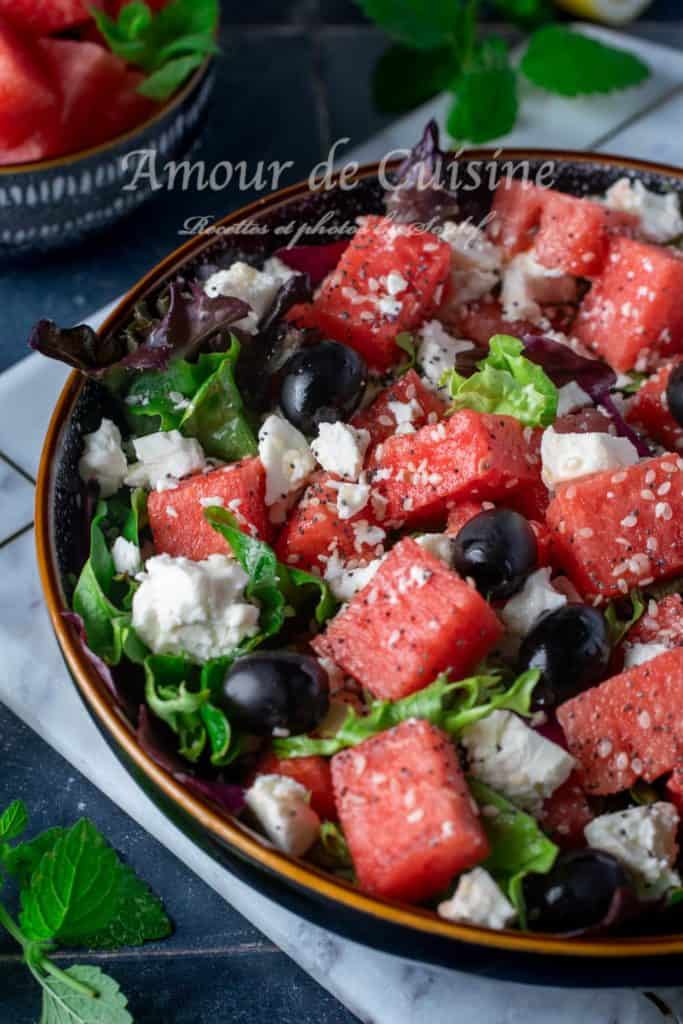 Close-up of a watermelon feta salad in a black bowl, featuring fresh lettuce, black olives, sesame seeds, and crumbled cheese, with mint leaves and watermelon pieces in the background