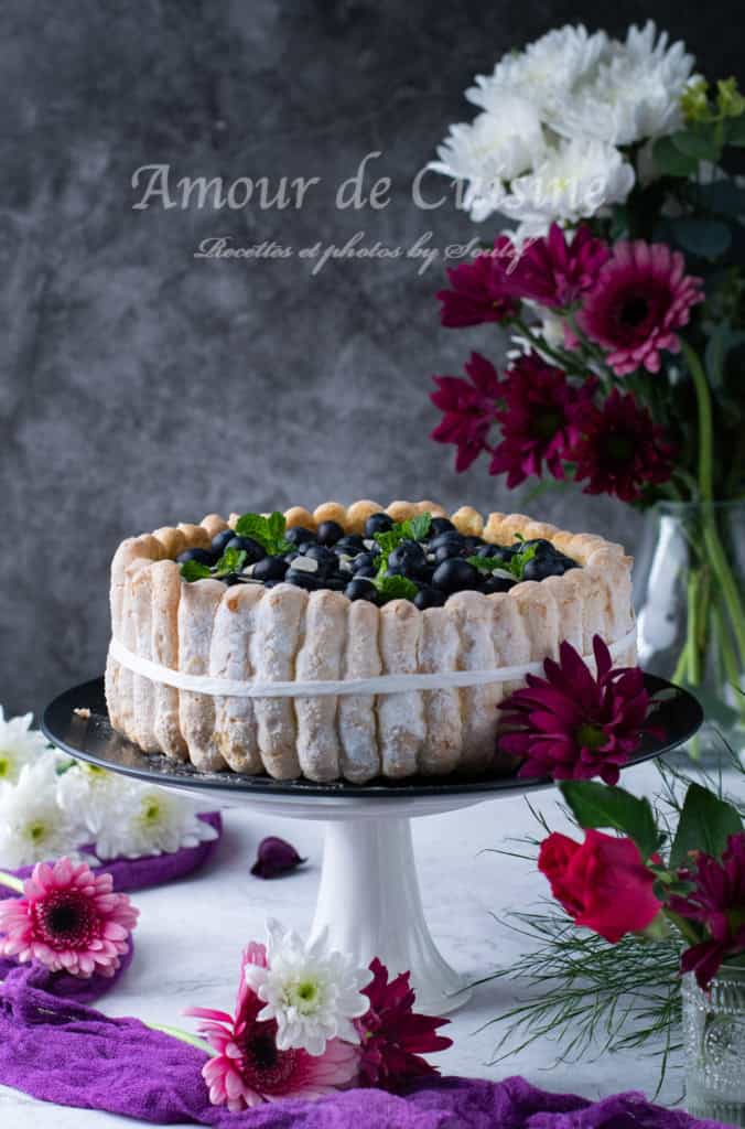 Blueberry charlotte cake on a stand, decorated with fresh blueberries, mint, and almond slices, surrounded by flowers.