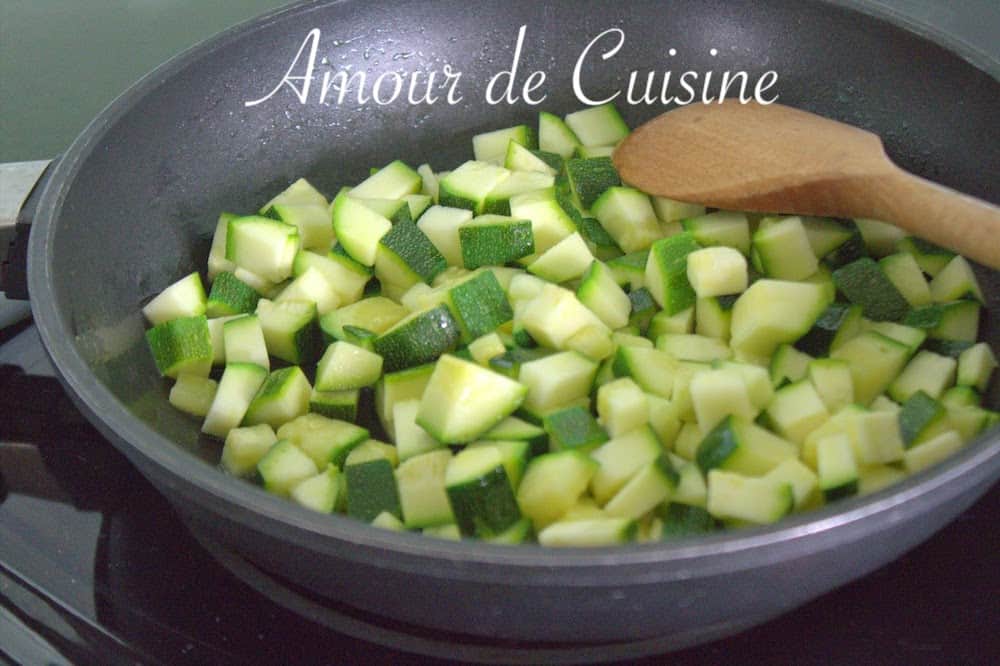 Diced zucchini being sautéed in a non-stick pan with a wooden spoon, part of the preparation for a savoury zucchini and feta cake