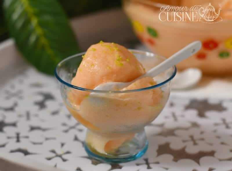 A glass cup filled with creamy homemade melon sorbet, topped with lime zest and served with a white spoon on a decorative tray, with a colorful bowl of sorbet in the background