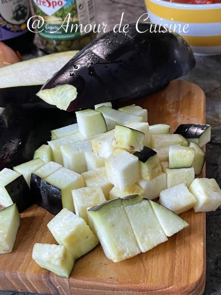 Fresh eggplant cut into cubes on a wooden cutting board, ready for cooking Moroccan zaalouk, with capers and tomato sauce jars in the background