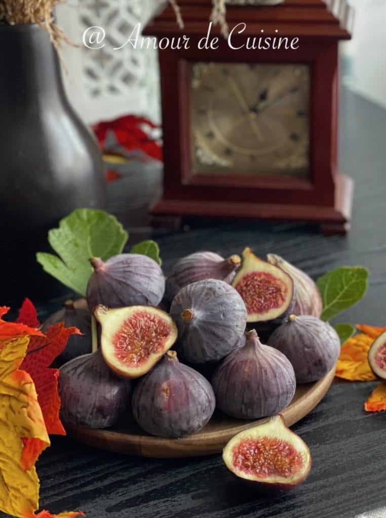 A rustic wooden plate filled with fresh purple figs, some whole and some cut open to reveal their rich, red interiors. The figs are garnished with green leaves and surrounded by vibrant autumn leaves. In the background, a vintage-style wooden clock and a dark vase add a cozy, seasonal ambiance.