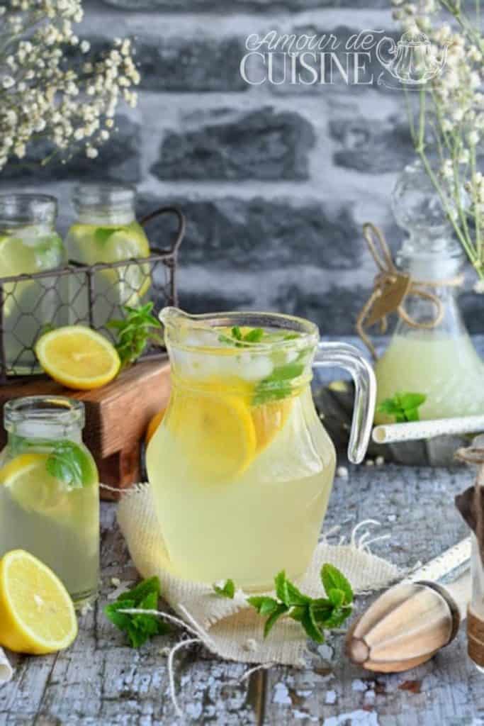Carafe de citronnade maison garnie de rondelles de citron et de feuilles de menthe fraîche, entourée de petits bocaux vintage, d’un demi-citron, de menthe fraîche et d’un presse-agrumes en bois, le tout posé sur une table rustique en bois vieilli avec un fond en pierre grise