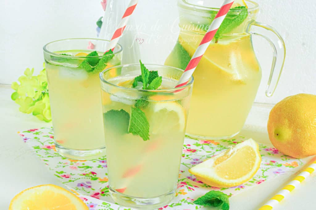 Chilled glasses of homemade lemonade with lemon slices, fresh mint, and striped straws, placed on a floral tray beside a glass pitcher of lemonade, surrounded by fresh lemons and mint leaves on a white table