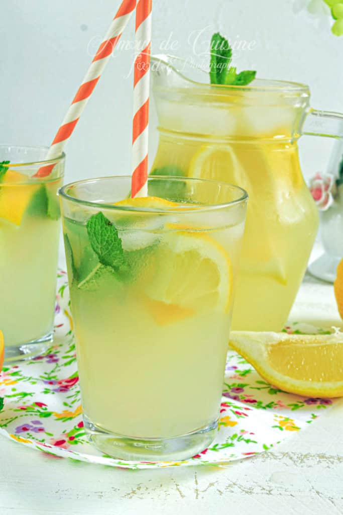 Glass of homemade lemonade with lemon slices, fresh mint, and ice, served with a red and white striped straw, next to a pitcher of lemonade and lemon wedges on a floral tray
