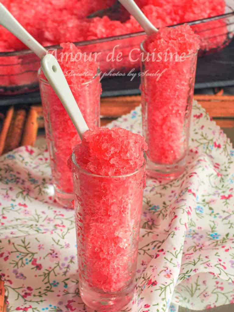Three tall glasses filled with bright red grenadine granita, each topped with a ceramic spoon, set on a floral cloth with a glass tray of granita in the background—refreshing homemade pomegranate ice treat.