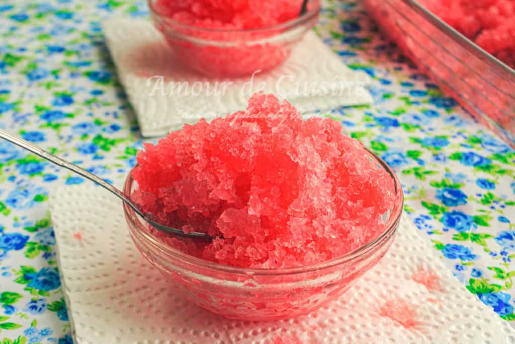 Close-up of a glass bowl filled with bright red grenadine granita, served with a spoon on a floral tablecloth—refreshing homemade frozen dessert made with pomegranate syrup.