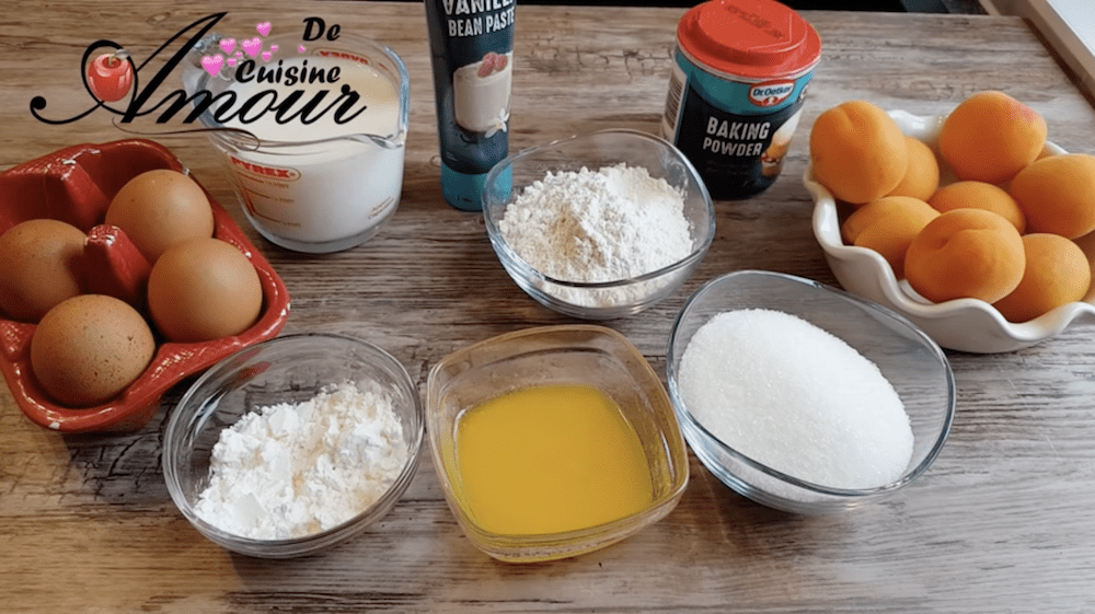 A top-down view of ingredients arranged on a wooden surface for making apricot clafoutis. The ingredients include fresh apricots in a white bowl, four brown eggs in a red ceramic holder, a measuring cup of milk, a tube of vanilla bean paste, a container of baking powder, bowls of flour, cornstarch, melted butter, and granulated sugar. The setup is neat and ready for baking, with the logo "De Cuisine Amour" displayed in the top left corner